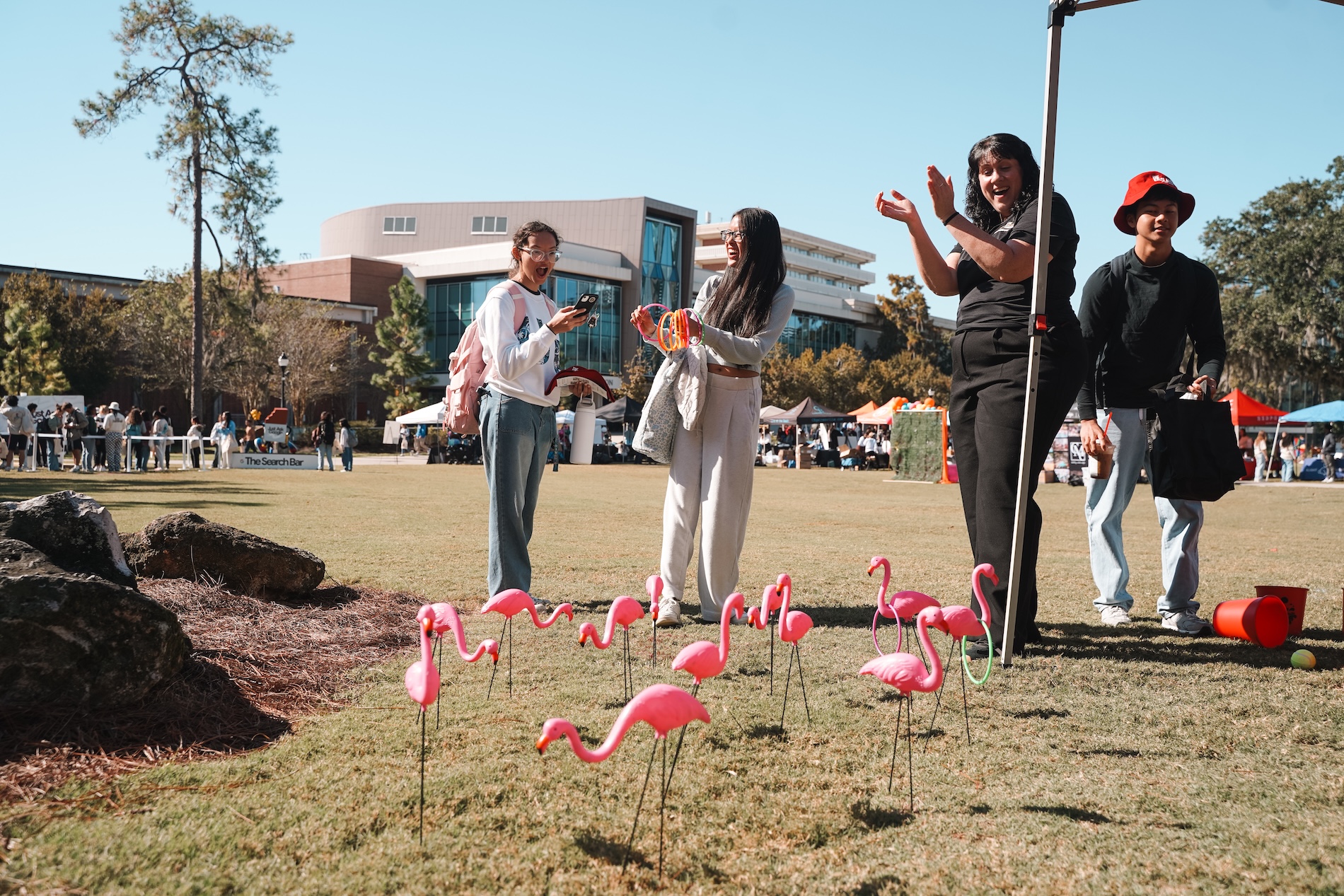 Two students tossing rings onto plastic flamingos. One student in shock and the other laughing, off-campus housing staff clapping, student in background with red hat on.